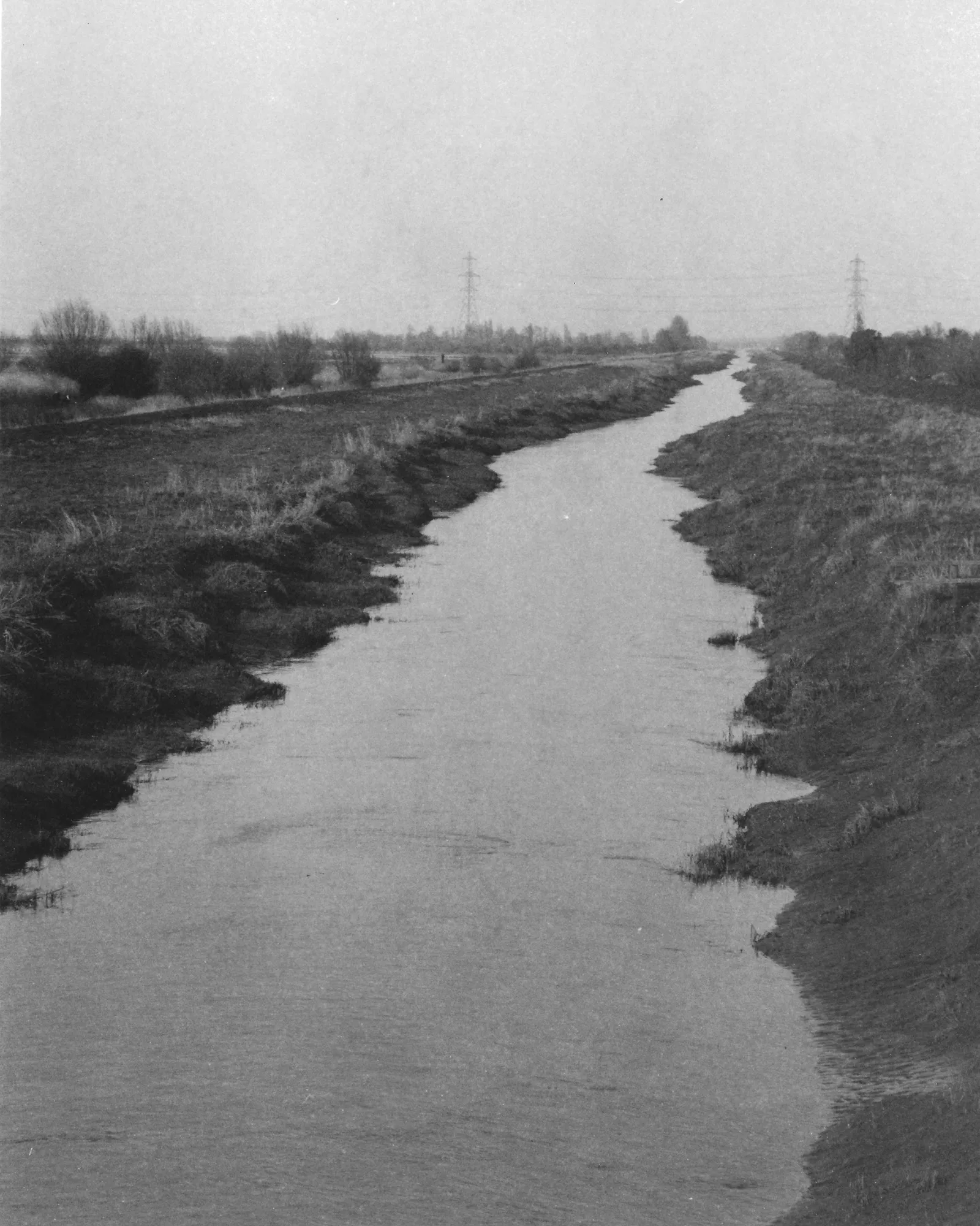 A view of New Bedford River from WWT Welney footbridge receding toward the horizon between the wetland reserve, flat farmland and two pylons in the distance